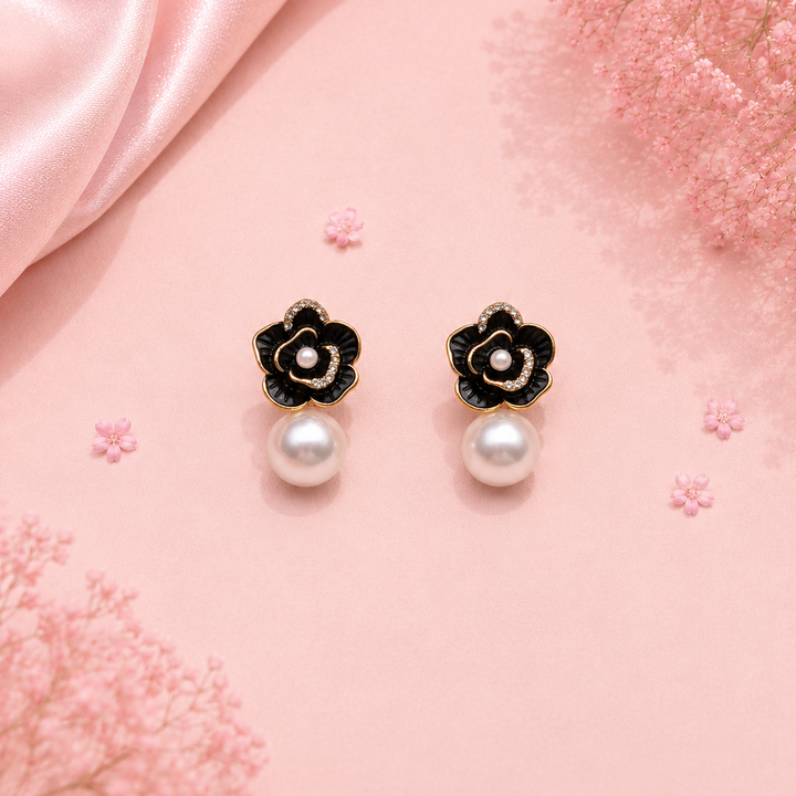 A pair of black and gold flower-shaped drop earrings with white pearl centers and hanging pearls, displayed on a pink satin background with decorative pink flowers.