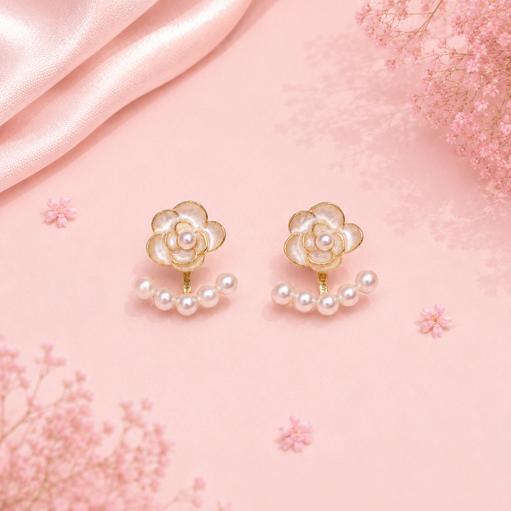 A pair of gold and pearl flower stud earrings displayed on a soft pink background with decorative pink satin fabric and dried baby's breath flowers.